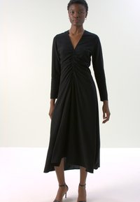 Vestido negro de manga larga con un diseño fruncido en el frente, dobladillo asimétrico, tela suave y escote en V. Combinado con zapatos de tacón alto.