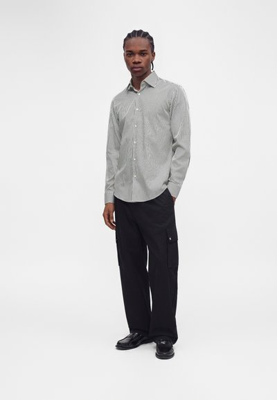 Homme debout portant une chemise blanche à manches longues avec de fines rayures noires, un pantalon cargo noir et des chaussures noires sur un fond blanc uni.