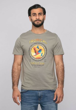 Homme portant un t-shirt gris avec des oiseaux tropicaux colorés et le texte "Tropical Sunset Bay", associé à un jean bleu, debout devant un fond uni.