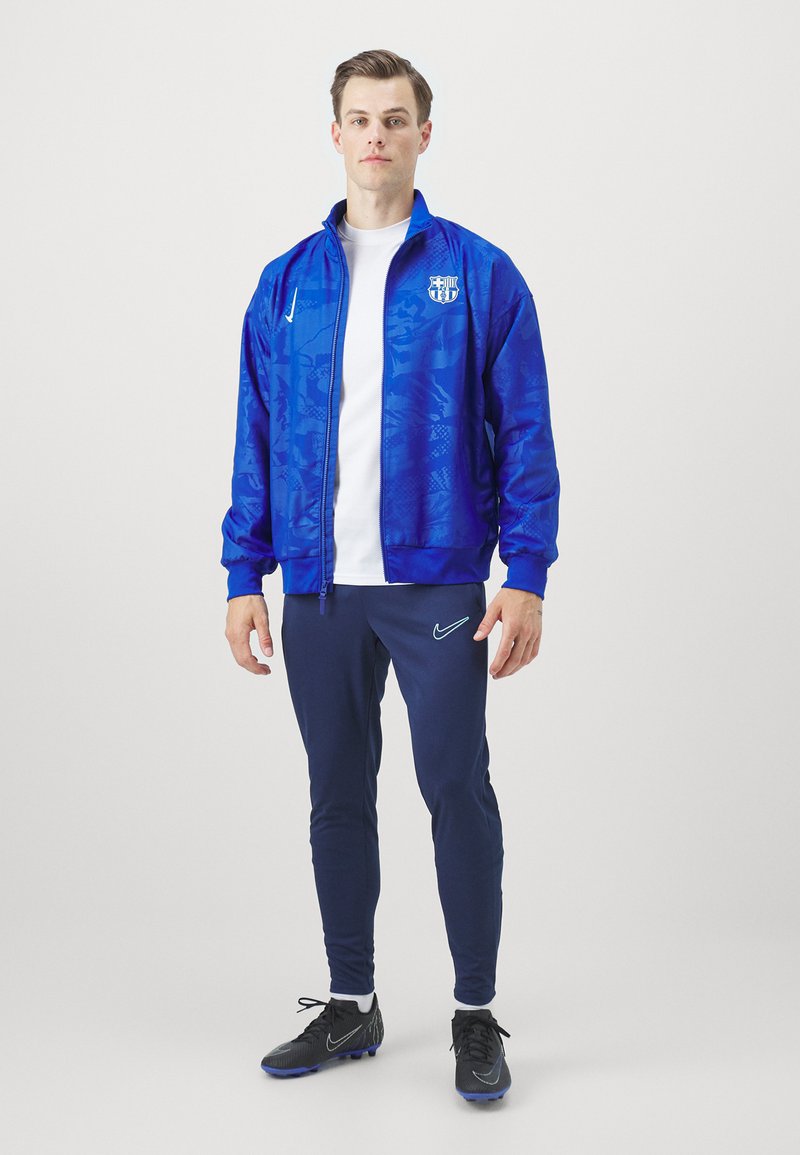 Chaqueta deportiva azul con acabado texturizado, camiseta blanca, pantalones azul marino y zapatillas deportivas negras. Presenta un logo en la chaqueta y los pantalones.