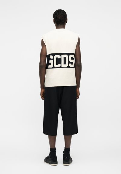 Άνδρας που στέκεται με την πλάτη στραμμένη, φοράει λευκό αμάνικο τοπ με μαύρη ταινία «GCDS», μαύρο σορτς με φαρδιά μπατζάκια, μαύρες κάλτσες και μαύρα αθλητικά παπούτσια.