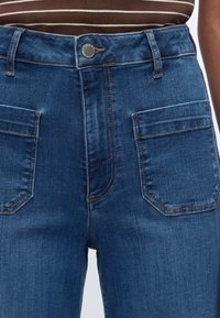Jean en denim bleu taille haute avec poches plaquées à l'avant, fermeture par boutons et coutures marron, porté avec un top rayé marron et blanc.