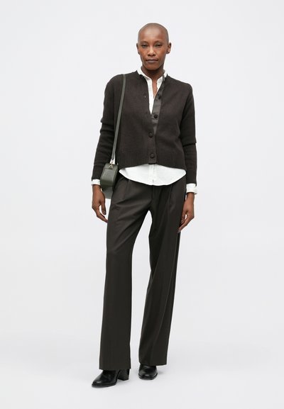 Bald woman wearing a dark cardigan, white shirt, wide dark trousers, black ankle boots, and carrying a dark shoulder bag, standing against a white background.