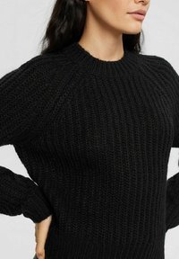 Svart stickad tröja med ribbat mönster, rund halsringning och raglanärmar, som visar en lös passform och ett mjukt, tjockt material.