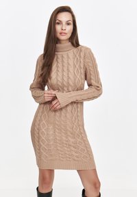 Beige Strickkleid mit hohem Kragen, das ein dickes Kabelmuster, lange Ärmel und gerippte Säume aufweist. Die Textur wirkt weich und gemütlich.