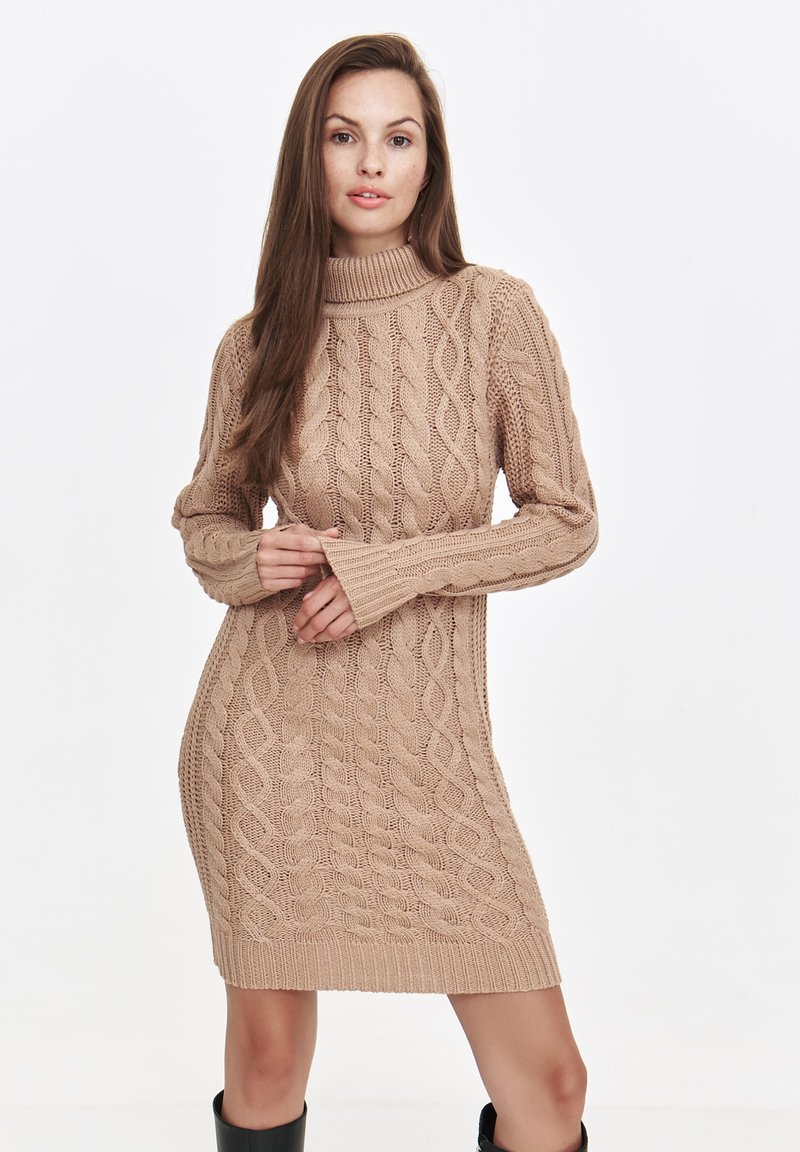 Beige Strickkleid mit hohem Kragen, das ein dickes Kabelmuster, lange Ärmel und gerippte Säume aufweist. Die Textur wirkt weich und gemütlich.