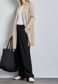 Béžový cardigan, bílé tričko, černé kalhoty s širokými nohavicemi a černá taška. Neformální tenisky s krémovými detaily. Jednoduché, čisté linie, měkké textury.