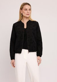 Czarna kurtka bomberka z teksturowanej tkaniny, z prążkowanymi mankietami i zamkiem błyskawicznym z przodu. Noszona na czarnym topie, w połączeniu z białymi spodniami.