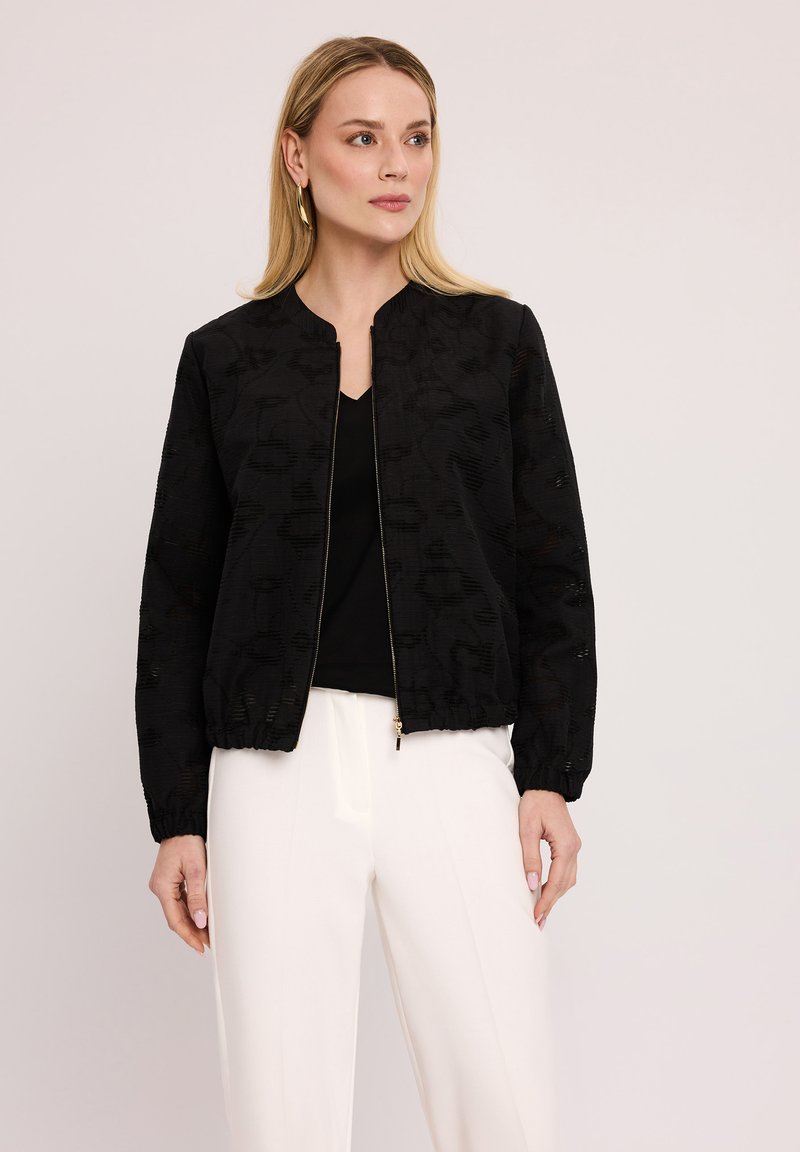 Czarna kurtka bomberka z teksturowanej tkaniny, z prążkowanymi mankietami i zamkiem błyskawicznym z przodu. Noszona na czarnym topie, w połączeniu z białymi spodniami.