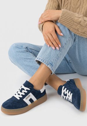 Osoba nosící modré semišové tenisky s bílými detaily, světle modré džíny a béžový svetr s copánkovým vzorem, sedí se zkříženýma nohama.