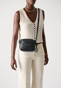 Schwarze Leder-Umhängetasche mit eingeprägtem Logo, rechteckige Form, dünner Schulterriemen, Details auf der Vorderseite, getragen über einem cremefarbenen Stricktop und hellen Hosen.