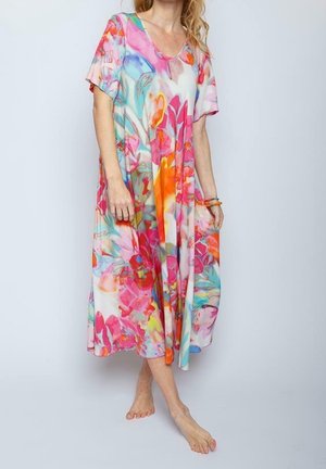 Kvinde stående barfodet i en løs, farverig midi-kjole med blomsterprint og korte ærmer mod en ensartet lys baggrund.