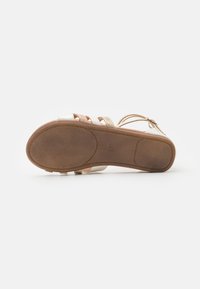 Flat sandal med en brun gummisåle og hvite stropper, med metalliske detaljer. Designet inkluderer flere vevde seksjoner for ventilasjon.