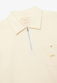 Polo in crema con colletto con zip, una tasca con accessori dorati e un'etichetta con il marchio. Texture morbida e dettagli di design minimal.