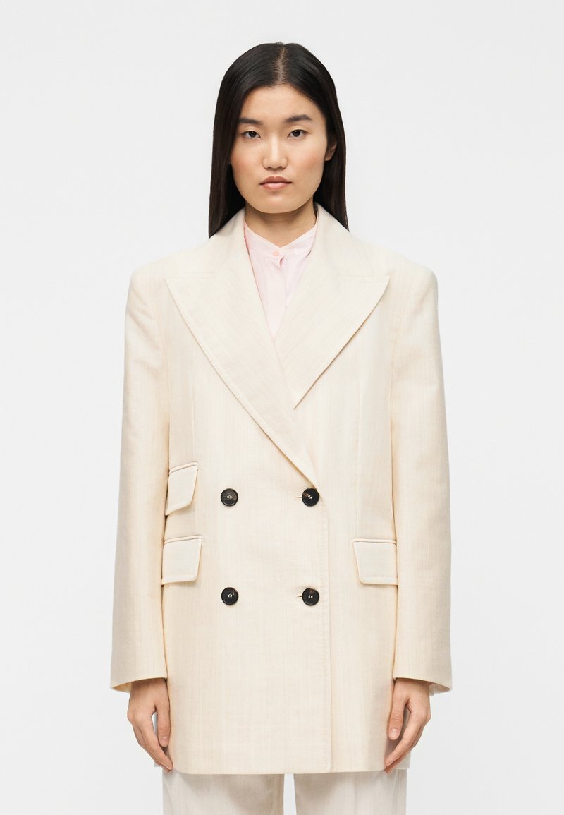 Μπεζ διπλής όψης σακάκι με μαύρα κουμπιά, μεγάλα πέτα και δύο τσέπες με καπάκι· κατασκευασμένο από υφασμάτινο ύφασμα με υφή.