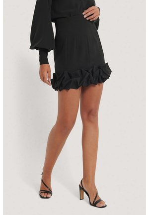 Femme portant une mini-jupe noire à ourlet volanté et des sandales noires à talons hauts, debout devant un fond uni.