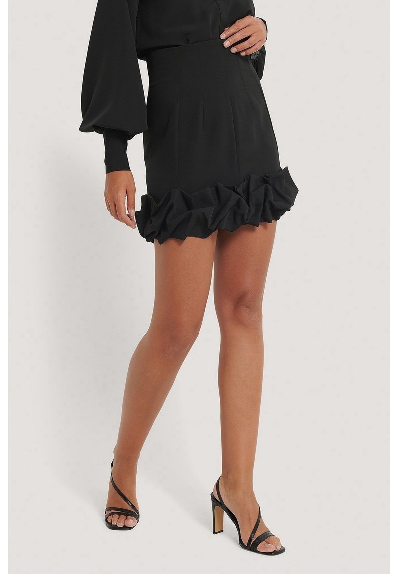 Femme portant une mini-jupe noire à ourlet volanté et des sandales noires à talons hauts, debout devant un fond uni.