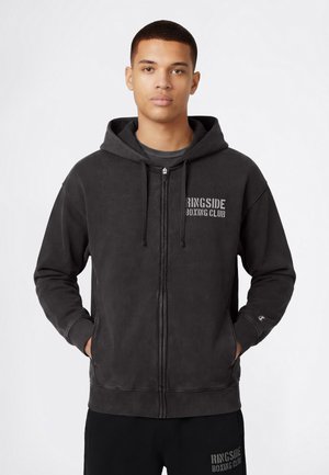 Jonge man draagt een zwart vest met ritssluiting en de tekst "Ringside Boxing Club", met handen in de zakken, staand tegen een effen witte achtergrond.