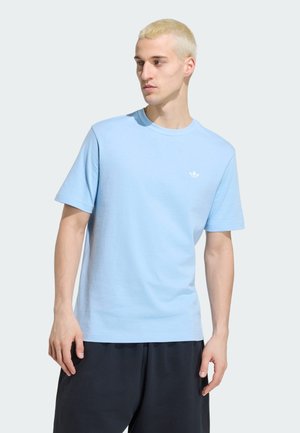 Junger Mann mit kurzen blonden Haaren, der ein hellblaues Adidas T-Shirt und schwarze Hose trägt, sieht nach links vor einem einfarbigen Hintergrund.