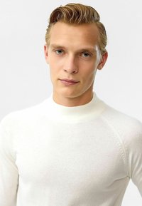 Witte gebreide trui met een hoge opstaande kraag, aansluitend ontwerp en raglanmouwen. Zachte textuur, minimale details en een gladde afwerking.