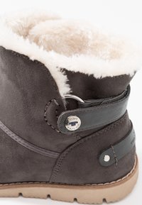 Tummanharmaat mokkasiinit talvikengät, joissa on vaaleanbeige vuori, koristeellinen nahkahihna, metallinen nappi ja vaaleanruskea kumipohja.