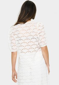 Top bianco in pizzo con un motivo a onde, maniche a sbuffo corte e vestibilità ampia, caratterizzato da una superficie testurizzata e un design trasparente.