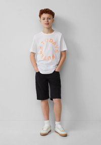 Weißes Baumwoll-T-Shirt mit orangefarbenem, kreisförmigem Text "HEUTE'S ZUKUNFT", kombiniert mit schwarzen Shorts und weißen Turnschuhen mit beigen Sohlen.