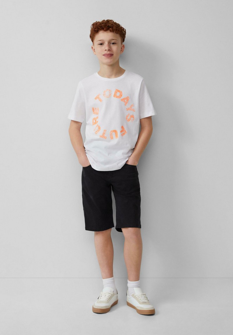 Weißes Baumwoll-T-Shirt mit orangefarbenem, kreisförmigem Text "HEUTE'S ZUKUNFT", kombiniert mit schwarzen Shorts und weißen Turnschuhen mit beigen Sohlen.