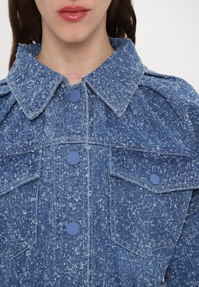 Jachetă din denim albastru cu un finisaj texturat, având un guler mare, butoane albastre și două buzunare pe piept. Țesătura prezintă un model cu pete.