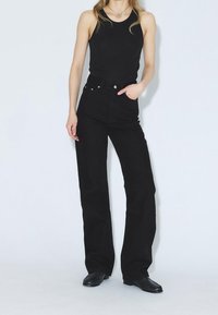 Vaqueros acampanados de mezclilla negra con cintura alta, diseño de cinco bolsillos y detalles de remaches de cobre. Combinados con una blusa negra sin mangas.