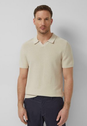 Mann trägt beigefarbenes kurzärmliges Strick-Poloshirt und dunkelblaue Hose, steht vor einem schlichten hellgrauen Hintergrund.