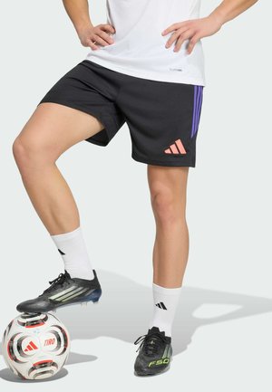 Personne portant un short de sport noir, des chaussettes blanches et des crampons noirs, équilibrant un ballon de football Adidas blanc et rouge sous un pied.