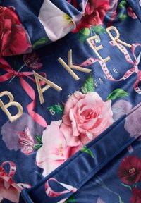 Tessuto in peluche blu navy con grandi motivi floreali colorati. Testo ricamato in oro che legge "BAKER" con nastri rosa sottostanti.