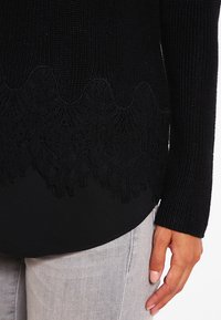 Svart stickad tröja med en scalloped spetskant. Har ett texturerat mönster och figurriktiga ärmar, i kombination med grå denim.