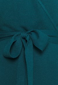Indumento lavorato a maglia color teal con texture a costine, caratterizzato da un design a incrocio e una cinturina annodata in vita. Superficie liscia con cuciture uniformi.