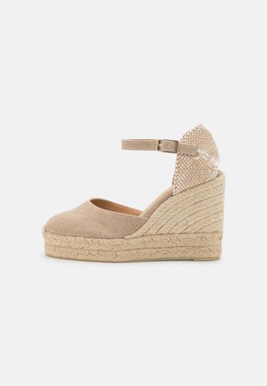 Beige kileespadrillesandal med ankelrem, flettet hæl, lukket tå og stofspænde på hvid baggrund.