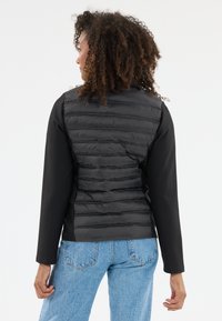 Svart pufferväst med quiltad textur och långa ärmar, i kombination med ljusblå jeans. Materialet har en slät yta.