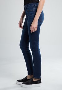Jeans ajustados de denim azul oscuro, con una cintura alta y una textura suave. Combinados con zapatos negros sin cordones con suela blanca.