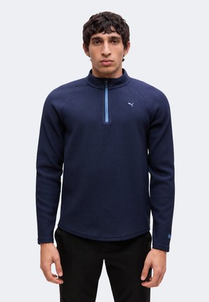 Man draagt een marineblauwe trui met lange mouwen, een vierdelige rits en een klein logo op de linkerborst tegen een effen achtergrond.