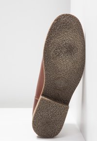 Zapato de cuero marrón con suela de goma texturizada. La suela presenta ligeros patrones de tracción y un detalle de logo grabado cerca del tacón.