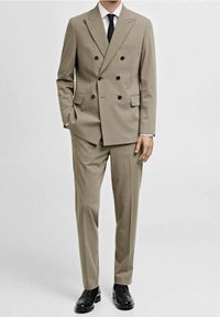 Uomo che indossa un completo beige doppiopetto con bottoni neri, camicia bianca, cravatta nera e scarpe eleganti nere, in piedi con una mano in tasca.