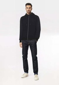 Felpa nera con zip e coulisse, abbinata a jeans in denim blu scuro e sneakers avorio. Design semplice con polsini a coste.