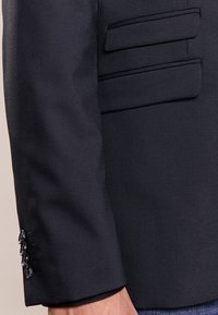 Dark navy blazer featuring a smooth texture, slim fit, and detailed flap pockets. Button accents are visible on the cuff.