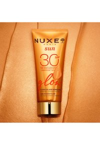 Fluide solaire éclat NUXE Sun SPF 30 avec protection anti-âge cellulaire, dans un tube orange sur une surface texturée bronze.