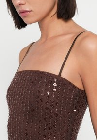 Καφέ φόρεμα διακοσμημένο με παγιέτες, με λεπτές τιράντες και εφαρμοστή κορυφή. Η υφή είναι λεία με παγιέτες σε σχέδιο λουλουδιών σε όλο το ύφασμα.