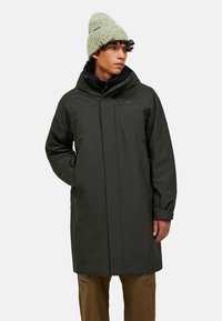 Tânăr îmbrăcat cu o caciulă verde și o haină de iarnă cu glugă de culoare verde închis, cu mâinile în buzunare, stând pe un fundal simplu deschis.