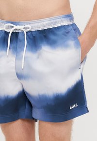 Mėlyni ir balti kaklaraištinio dažymo plaukimo šortai su elastinga juosmens juosta, raišteliu, šoninėmis kišenėmis ir mažu baltu logotipu apačioje.