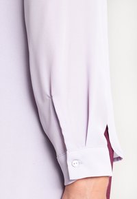 Blusa de color púrpura claro con mangas de textura suave, que presenta un puño abotonado y una sutil abertura lateral para el ajuste de la talla.
