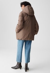 Giacca a vento marrone con cappuccio, caratterizzata da un esterno liscio e una vestibilità rilassata, abbinata a jeans cropped azzurri chiari e stivali neri alla caviglia.