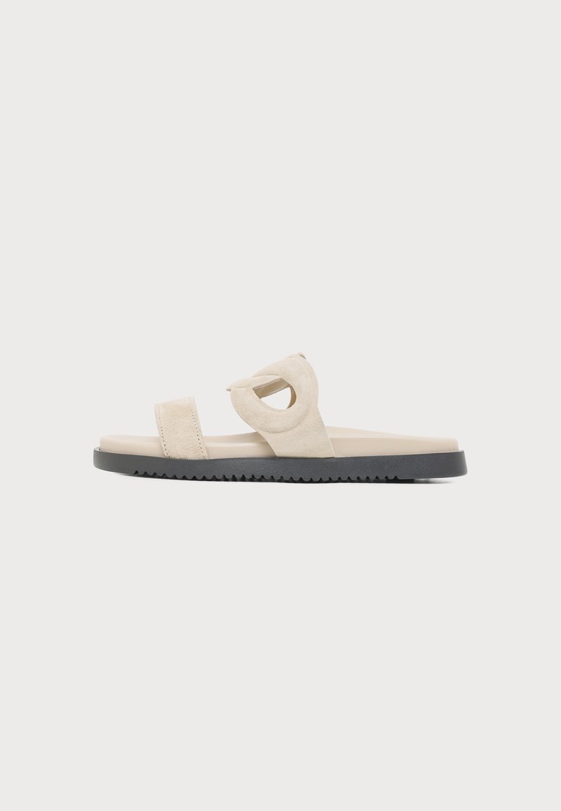 Sandale beige à enfiler avec deux larges brides, dont une avec une découpe ronde, et une semelle noire texturée sur un fond uni.